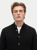 A man wearing a black cable-knit button-up cardigan sweater over a white t-shirt, photographed against a solid light gray background.
