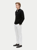 A man wearing a black cable-knit button-up cardigan over a white collared shirt, paired with white trousers and black leather loafers, standing in profile against a light gray studio background.