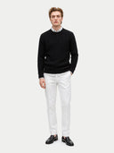 A man modeling a black cable-knit crewneck sweater over a white collared shirt, paired with white trousers and black leather loafers, against a light gray studio background.