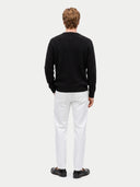 A man viewed from behind wearing a black cable-knit sweater, white trousers, and black loafers, standing against a light gray studio background.