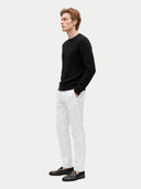A man in a black cable-knit crewneck sweater, white trousers, and black loafers, standing in profile against a light gray studio background.
