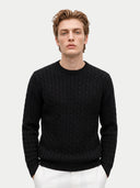 A man wearing a black cable-knit crewneck sweater and white trousers, standing against a light gray studio background.