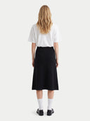 A woman is shown from behind wearing a white short-sleeve t-shirt, a black midi skirt, white socks, and black loafers, against a light gray studio background.
