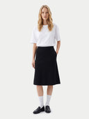 A woman stands against a light gray studio background wearing a white short-sleeve crewneck T-shirt, a black knee-length A-line skirt, white knee-high socks, and black leather loafers.