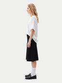 A woman stands in profile wearing an oversized white short-sleeve top, a black midi skirt, white socks, and black loafers against a plain light gray studio background.