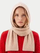 A woman wearing a ribbed knit hooded scarf in a light beige color, styled over a bright red cable-knit sweater against a light gray background.
