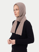 A woman wearing a ribbed taupe hijab and a black cable-knit sweater, shown from the chest up against a light gray studio background.