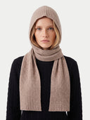 A model wearing a ribbed knit hooded beanie and a matching long scarf in a light taupe-beige color, paired with a black cable-knit sweater against a plain light gray background.