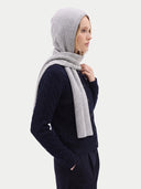 A woman shown in profile wearing a light gray ribbed knit headscarf and matching scarf wrapped around her shoulders, over a dark navy cable-knit cardigan and navy pants, against a plain light gray studio background.