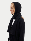 A woman in profile wearing a black ribbed knit hood and matching scarf over a black cardigan, against a light gray studio background.