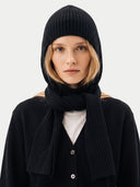 A model wearing a black ribbed knit hooded beanie, a matching black scarf, and a black button-front cardigan over a light gray top, photographed against a plain light gray background.
