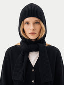 A model wearing a black ribbed knit hooded beanie, matching scarf, and black cardigan over a light gray shirt, photographed against a plain light gray background.