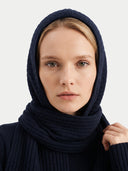 A close-up of a woman wearing a dark navy blue ribbed knit hood and matching scarf against a light gray background.