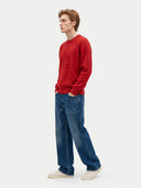 A man wearing a red crewneck sweatshirt with a tonal embroidered graphic on the chest, paired with wide-leg blue jeans and off-white sneakers, standing against a light gray studio background.