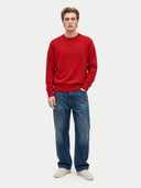 A male model stands against a light gray studio background wearing a bright red crewneck sweatshirt with a tonal embroidered logo on the chest, blue jeans, and off-white sneakers.