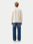 A man viewed from behind wearing a cream-colored crewneck sweater and blue jeans against a light gray studio background.