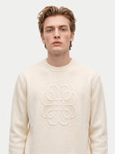 A man wearing a cream-colored knit sweater with a large tonal embroidered Loewe logo on the chest, set against a plain light gray background.