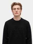 A man wearing a black crewneck sweater with a tonal, raised geometric logo on the chest, photographed against a light gray studio background.