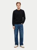 A man modeling a black crewneck sweater with a tonal embroidered logo on the chest, paired with blue jeans and beige sneakers, against a light gray studio background.