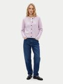 A woman stands against a light gray studio background wearing a light lavender cable-knit button-up cardigan over a white top, blue jeans, and black leather shoes.