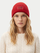 A woman with blonde hair wearing a red ribbed knit beanie with a black 'GOBI EST.1981' patch on the front, paired with a cream cable-knit sweater against a light gray background.