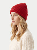 A woman in profile wearing a red ribbed knit beanie with a small black rectangular logo patch on the front, paired with a cream cable-knit sweater against a light gray background.