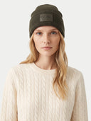 A woman wearing a dark olive green ribbed knit beanie with a rectangular patch that reads 'GOBI EST. 1981' and a cream cable-knit sweater, photographed against a light gray studio background.
