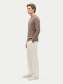 side view of a man standing wearing a taupe colored crew neckline cable sweater, white pants and sneakers on a light gray background.