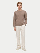 A male model wearing a taupe cable-knit crewneck sweater, off-white linen trousers, and light beige sneakers, standing against a solid light gray background.