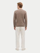 A man viewed from the back wearing a taupe cable-knit sweater and off-white trousers, standing against a light gray studio background.