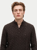 A man wearing a dark brown cable-knit polo sweater with a buttoned mock neck collar, photographed against a light gray studio background.