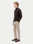 A man wearing a dark brown textured knit mock-neck sweater, beige trousers, and dark brown leather loafers, standing in profile against a light gray studio background.