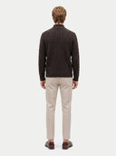 A man viewed from the back wearing a dark brown cable-knit sweater, beige trousers, and brown leather loafers, set against a plain light gray studio background.