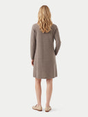 A woman viewed from the back wearing a long-sleeve, knee-length taupe knit dress and beige loafers, set against a plain light gray studio background.