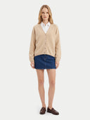 A woman stands against a light gray studio background wearing a beige button-front cardigan over a white collared shirt, a dark blue denim mini skirt, and brown leather loafers.
