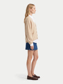 A woman shown in profile wearing a beige cardigan over a white collared shirt, blue denim shorts, and dark brown loafers, photographed against a clean light gray studio background.