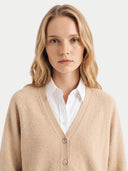 A woman with blonde hair wearing a beige knit cardigan over a white collared shirt, photographed against a clean light gray studio background.