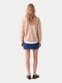 A woman viewed from behind wearing a beige knit sweater, a blue denim mini skirt, a white undershirt, and brown loafers, set against a plain light gray studio background.