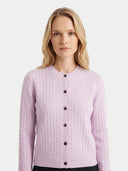 A woman with blonde hair wearing a light pink cable-knit cardigan sweater with dark buttons, photographed against a plain light gray studio background.