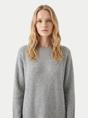 A blonde woman wearing a light gray knit crewneck sweater, photographed against a clean light gray studio background.