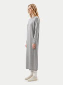 A woman modeling a light gray long-sleeve knit maxi dress, paired with white socks and beige sneakers, against a solid off-white studio background.