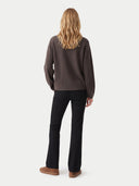 A woman is shown from the back wearing a dark brown knit sweater, black pants, and brown suede shoes against a light gray studio background.