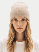 A woman with long blonde hair wearing a beige ribbed knit beanie with a tonal embroidered logo, paired with a white crewneck sweatshirt, against a light gray background.