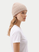 side view of a woman wearing a beige beanie with brand logo and white t-shirt on a light gray background.