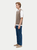 A man wearing a light brown cable-knit sweater vest over a white t-shirt, paired with blue jeans and beige shoes, standing against a light gray studio background.