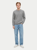 A man wearing a gray cable-knit crewneck sweater, light blue jeans, and white sneakers, standing against a light gray studio background.