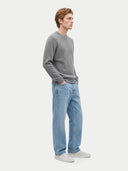 A man wearing a gray cable-knit crewneck sweater, light blue jeans, and white sneakers, standing in profile against a light gray studio background.