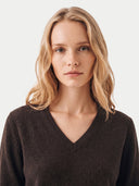 A blonde woman with a neutral expression wearing a dark brown V-neck knit sweater, photographed against a clean off-white background.