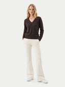 A woman modeling a dark brown V-neck long-sleeve sweater and cream-colored wide-leg trousers against a light gray studio background.