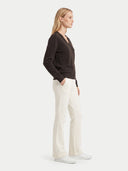 A woman in side profile wearing a dark brown V-neck knit sweater, off-white wide-leg trousers, and white sneakers, standing against a light gray studio background.